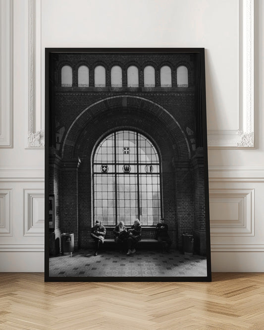 A dramatic black and white photograph of a large, brick-walled waiting area, possibly a train station. Four people are seated on a long bench beneath a massive arched window, which dominates the scene. The lighting creates a moody, contemplative atmosphere, highlighting the grandeur of the architecture. Artwork