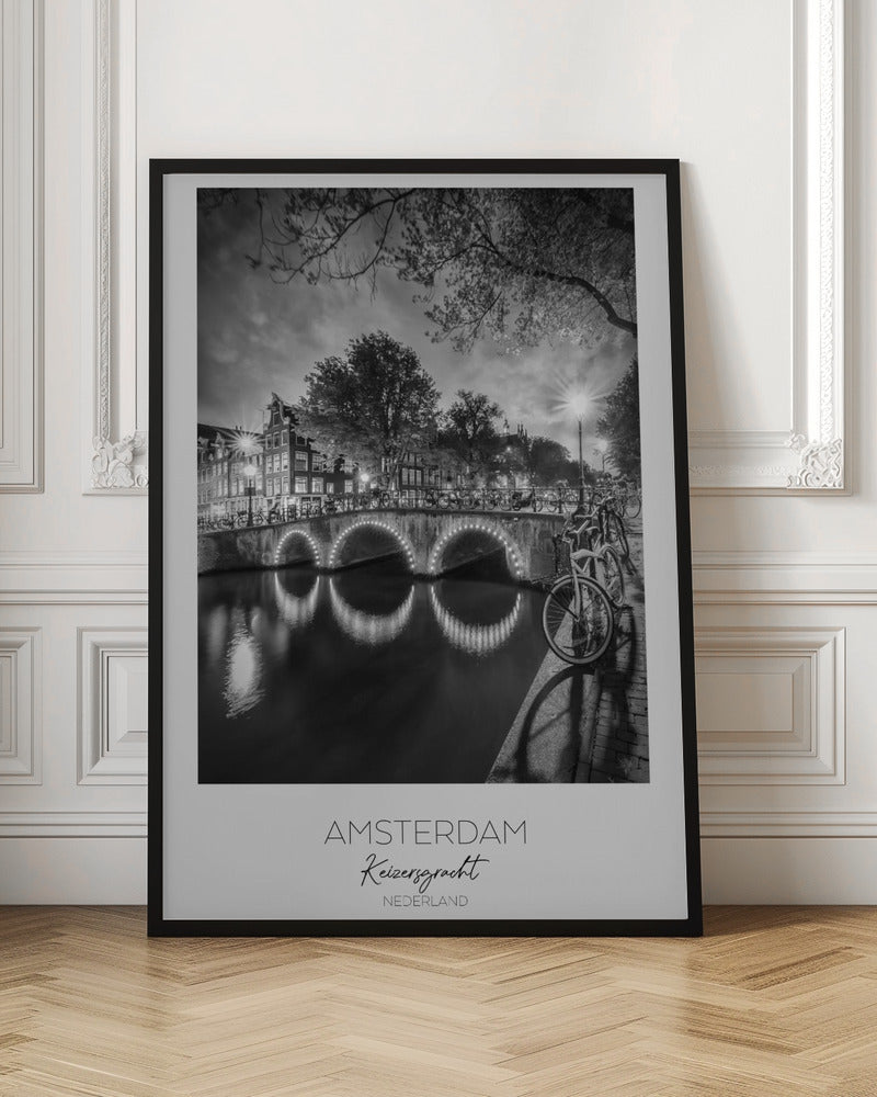 A black and white poster of Amsterdam's Keizersgracht canal at night. An arched bridge with glowing lights is the centerpiece, its reflection shimmering on the dark water. Bicycles are parked along the bridge and in the foreground on a cobblestone path. Classic canal houses and trees line the background under a dramatic, cloudy sky. The image is framed with text below reading 'AMSTERDAM Keizersgracht NEDERLAND'. Artwork
