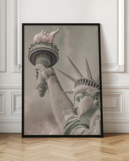 A low-angle, close-up photograph of the Statue of Liberty with a vintage color treatment. The shot captures her profile, crown, and her arm holding the torch aloft against a muted, cloudy sky, all enclosed in a thin black frame. Poster