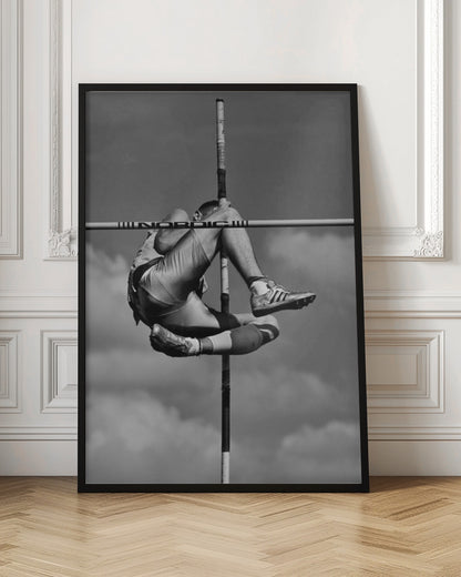 A dramatic black and white, low-angle photograph of a male athlete in mid-air during a pole vault. He is arching his back and bending his legs to clear the horizontal bar, with the vertical pole bisecting the frame. The background is a partly cloudy sky. Wall Art