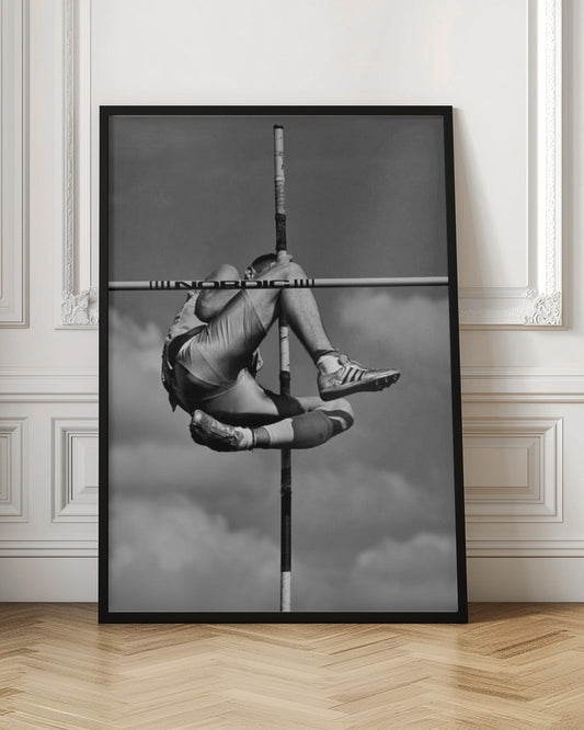 A dramatic black and white, low-angle photograph of a male athlete in mid-air during a pole vault. He is arching his back and bending his legs to clear the horizontal bar, with the vertical pole bisecting the frame. The background is a partly cloudy sky. Wall Art