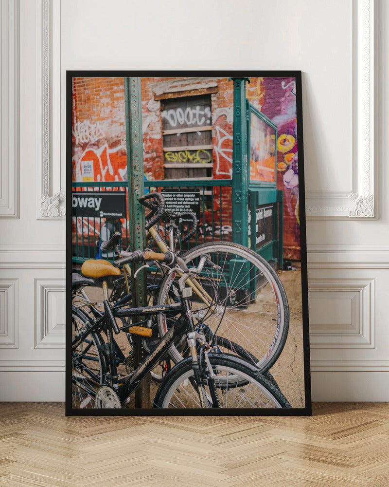 Several bicycles are locked to a green street pole in front of a red brick building covered in graffiti, with a city subway entrance visible in the background. Poster