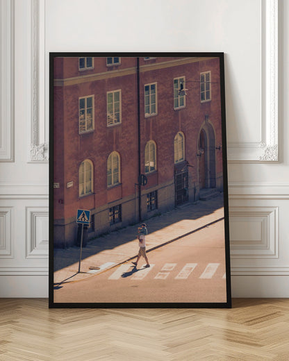 A high-angle photograph of a man carrying a child on his shoulders as they walk across a sunlit crosswalk, with a large, reddish-brown European-style building in the background. The scene is bathed in warm light, creating long shadows. Poster