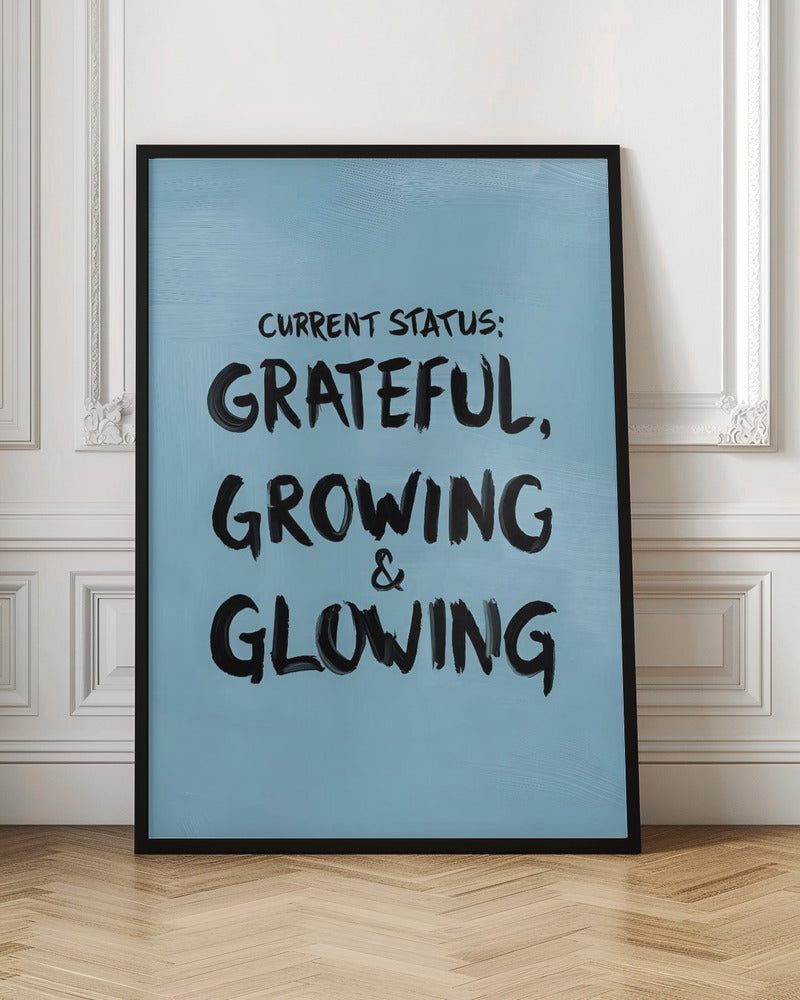 A motivational poster with a light blue background and a thin black frame. The text 'CURRENT STATUS: GRATEFUL, GROWING & GLOWING' is written in a bold, black, handwritten brush font. Decor