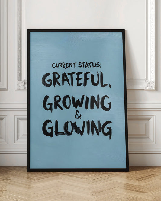 A motivational poster with a light blue background and a thin black frame. The text 'CURRENT STATUS: GRATEFUL, GROWING & GLOWING' is written in a bold, black, handwritten brush font. Decor