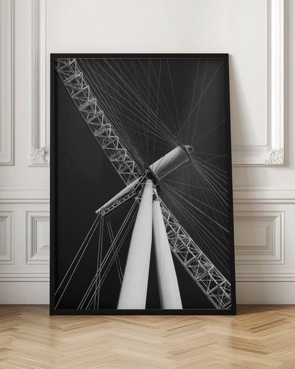 A dramatic low-angle, black and white photograph of a large Ferris wheel, focusing on the central hub and supporting structure. The image captures the white A-frame legs and the intricate lattice of the wheel's rim, with numerous tension cables radiating outwards against a solid black sky, creating a strong sense of perspective and geometric pattern. Artwork