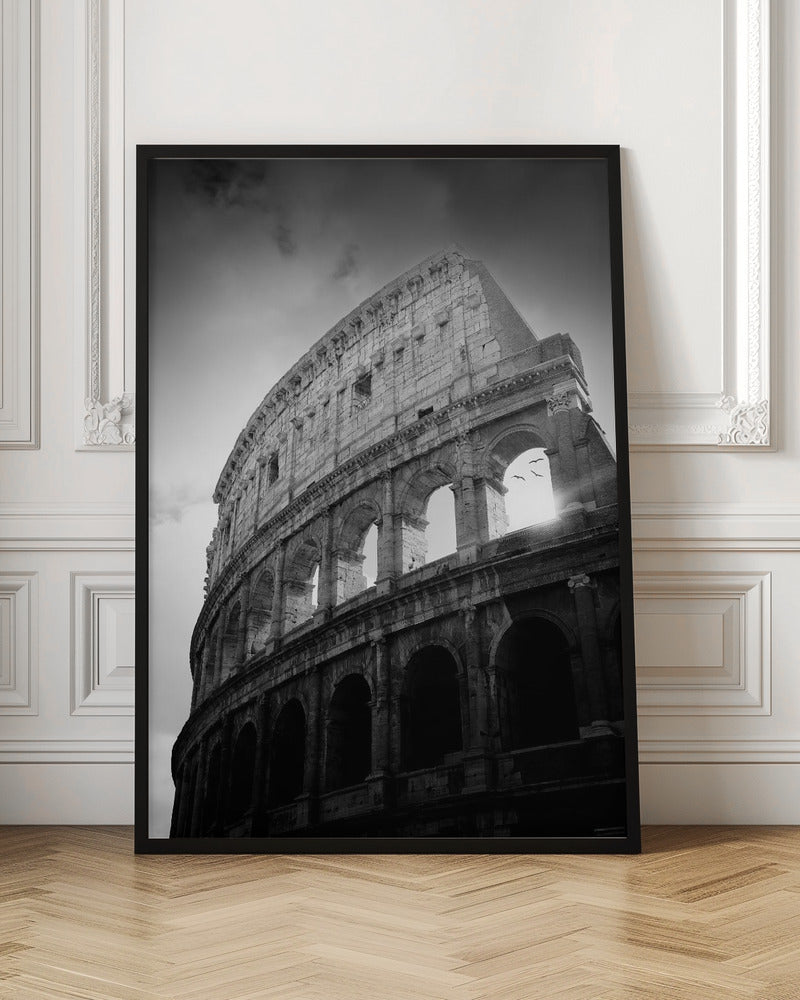 A dramatic, low-angle black and white photograph of the ancient Roman Colosseum. The sun shines brightly through one of the upper archways, creating a lens flare and silhouetting a few birds in flight against the cloudy sky. Poster