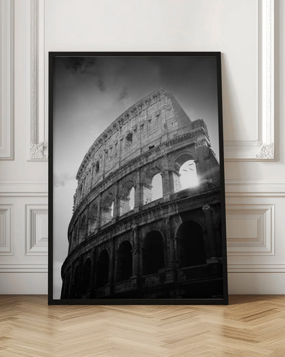 A dramatic, low-angle black and white photograph of the ancient Roman Colosseum. The sun shines brightly through one of the upper archways, creating a lens flare and silhouetting a few birds in flight against the cloudy sky. Poster