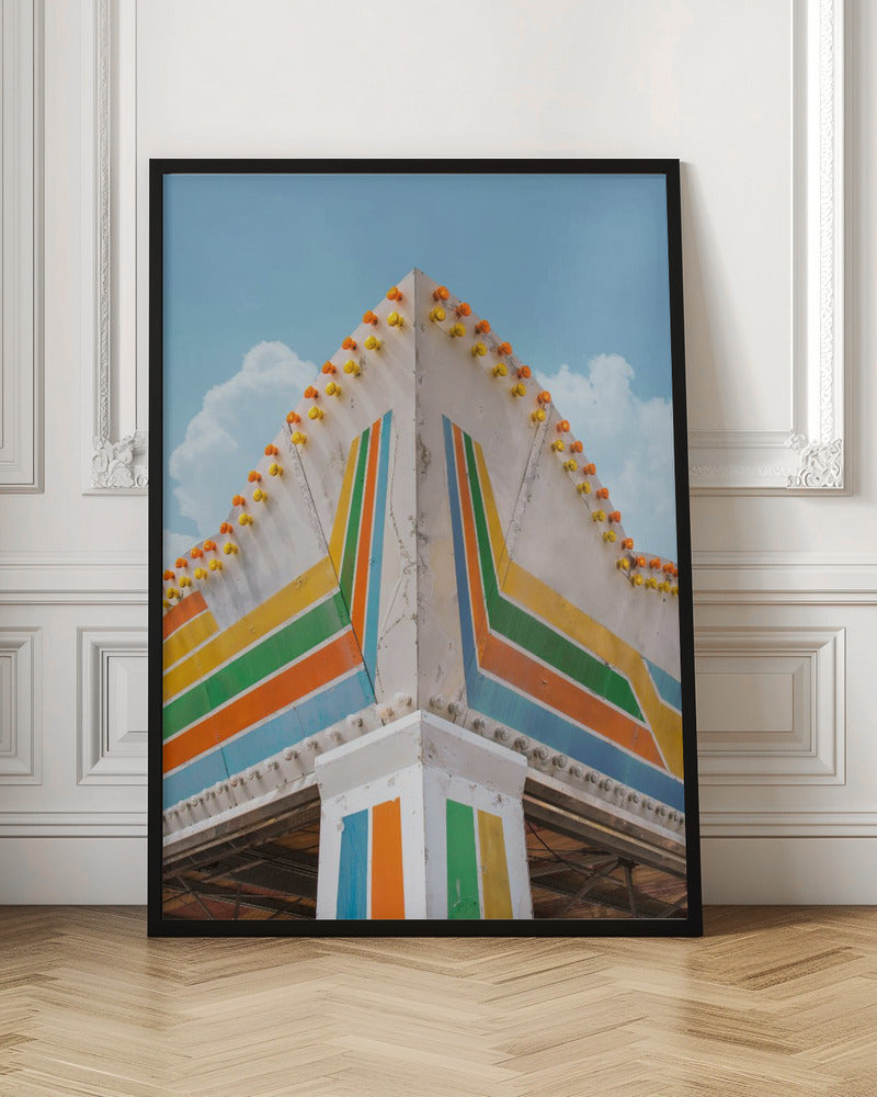 A low-angle photograph of the corner of a retro-style building against a bright blue sky with white clouds. The white facade is painted with bold, colorful stripes in orange, yellow, green, and blue, and the angled roofline is decorated with rows of unlit yellow and orange carnival lights. Print