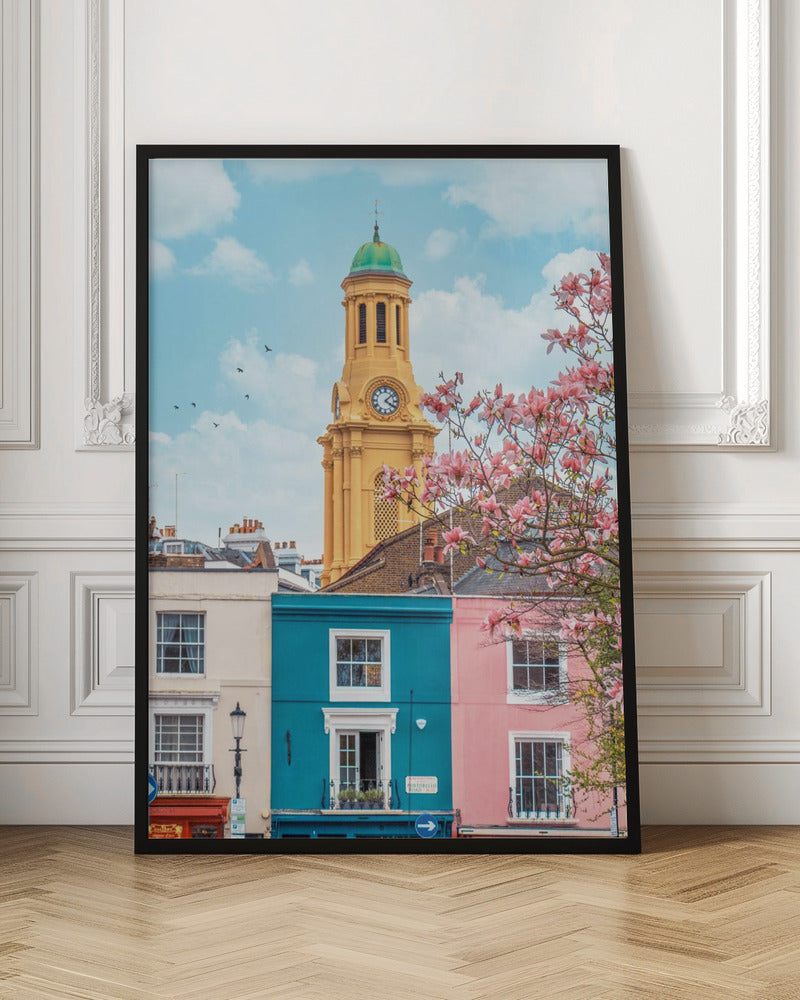 A vibrant city street scene on a sunny day, featuring a row of colorful houses in teal, pink, and off-white. A striking yellow clock tower with a green dome stands tall against a bright blue sky, with a blooming pink magnolia tree in the foreground. Print