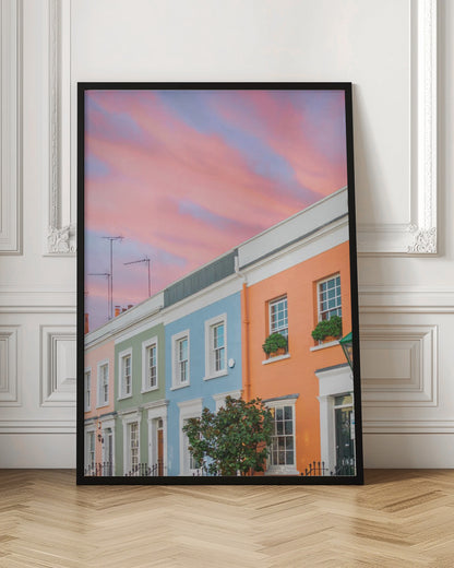 A row of colorful pastel townhouses in shades of orange, blue, green, and pink, viewed from a low angle against a dramatic, cloudy pink and purple sunset sky. Poster