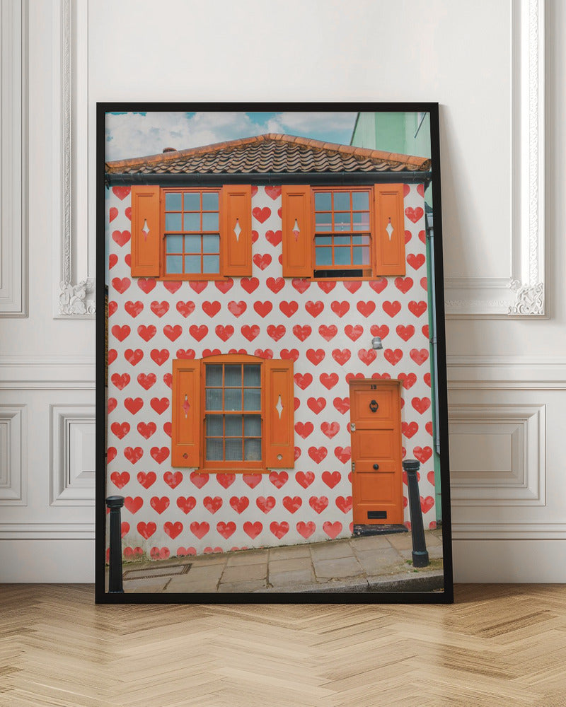 The front facade of a whimsical two-story house with white walls covered in a pattern of red hearts. The house has bright orange shutters on its windows and a matching orange front door, creating a vibrant and playful look against the heart-patterned background. Artwork