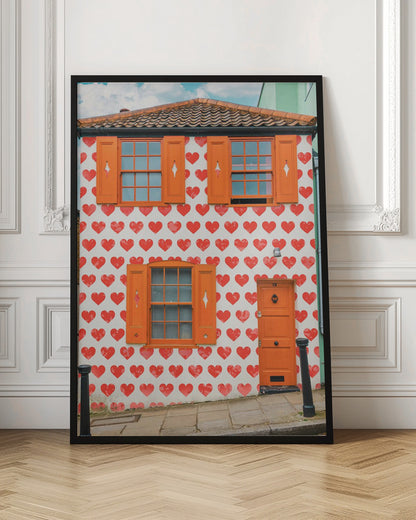 The front facade of a whimsical two-story house with white walls covered in a pattern of red hearts. The house has bright orange shutters on its windows and a matching orange front door, creating a vibrant and playful look against the heart-patterned background. Artwork
