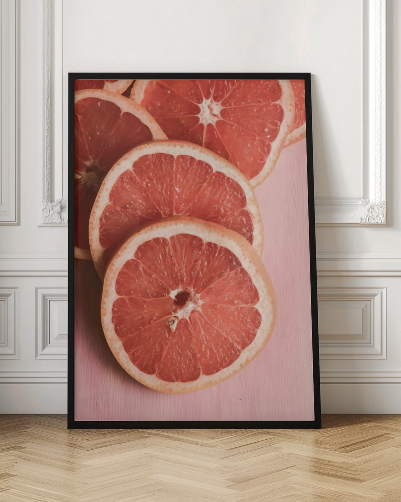 A close-up photograph of several overlapping slices of vibrant pink grapefruit arranged on a pale pink surface, enclosed within a simple black frame. Print