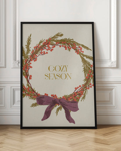 A vintage-style illustration of a festive wreath made of green boughs and red berries, tied with a large purple bow at the bottom. The text 'COZY SEASON' is written in gold inside the wreath on a cream-colored background, all enclosed in a thin black frame. Wall Art