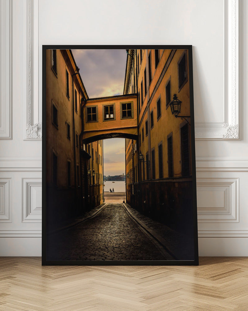 A perspective view down a narrow, shadowy cobblestone alley in an old European city, framed by warm, ochre-colored buildings. An arched walkway connects the buildings overhead, and the alley opens to a view of a tranquil body of water at sunset, with two small figures silhouetted by the shore. Decor