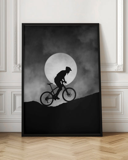 A dramatic black and white silhouette of a mountain biker riding uphill against the backdrop of a large, full moon in a cloudy night sky. Decor