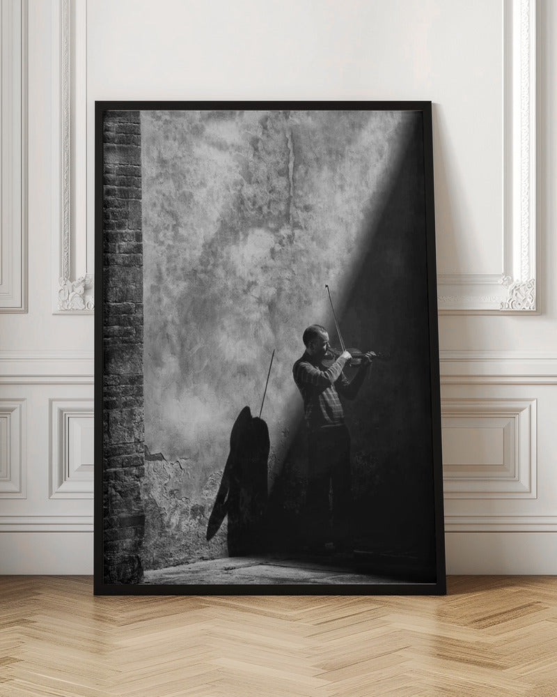 A dramatic black and white photograph of a man playing the violin, standing in a corner against a weathered wall. He is bathed in a sharp diagonal ray of light, casting a long, dark shadow behind him. Artwork