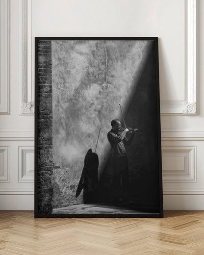 A dramatic black and white photograph of a man playing the violin, standing in a corner against a weathered wall. He is bathed in a sharp diagonal ray of light, casting a long, dark shadow behind him. Artwork