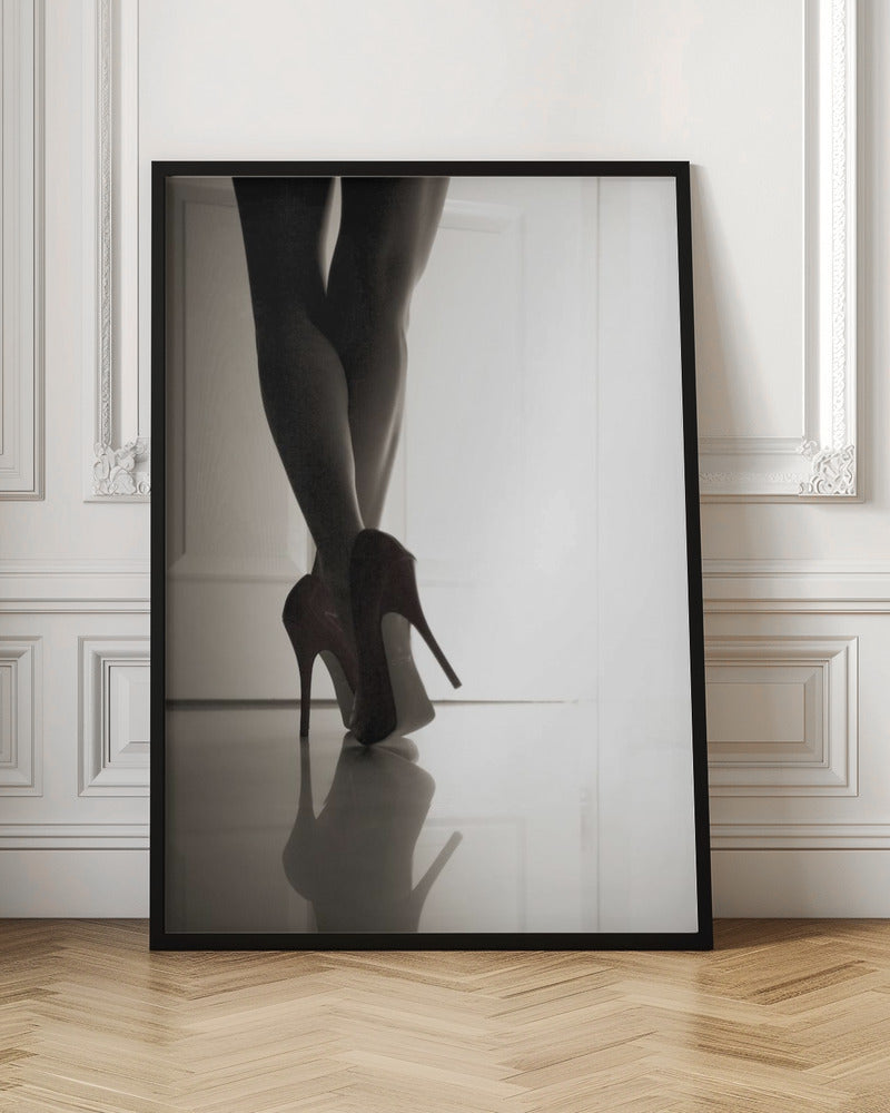 A low-angle, black and white photograph focusing on a woman's legs as she stands with them crossed. She wears dark red stiletto high heels, which cast a clear reflection on the glossy floor. Her legs are silhouetted against a bright white door in the background, creating a dramatic and sensual mood. Decor