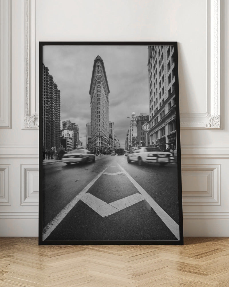 A dynamic black and white, low-angle photograph of the iconic Flatiron Building in New York City. The street in the foreground has bold white lines leading the eye to the building, while cars on either side are blurred with motion, capturing the city's fast pace under a cloudy sky. Decor