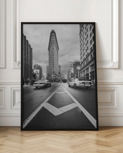 A dynamic black and white, low-angle photograph of the iconic Flatiron Building in New York City. The street in the foreground has bold white lines leading the eye to the building, while cars on either side are blurred with motion, capturing the city's fast pace under a cloudy sky. Decor