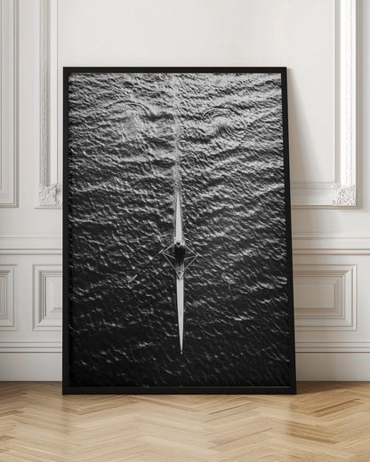 A dramatic black and white overhead photograph of a lone rower in a sleek racing shell, cutting a straight path through dark, textured water. The high-contrast lighting highlights the ripples on the water's surface and the rower's focused form. Artwork