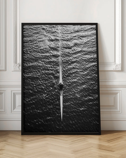 A dramatic black and white overhead photograph of a lone rower in a sleek racing shell, cutting a straight path through dark, textured water. The high-contrast lighting highlights the ripples on the water's surface and the rower's focused form. Artwork