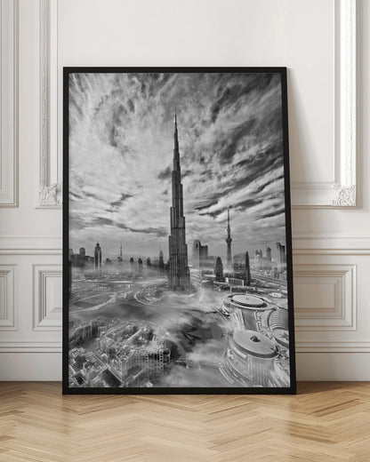 A dramatic black and white high-angle photograph of the Burj Khalifa dominating the Dubai cityscape, with swirling clouds above and a light mist surrounding the buildings below, all enclosed in a black frame. Poster