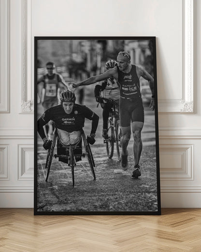 A dynamic black and white action photograph of a para-athlete in a racing wheelchair during a race. A running athlete beside him reaches out to hand him a sponge, showcasing a moment of camaraderie and support. Other competitors are blurred in the background on the wet asphalt. Print