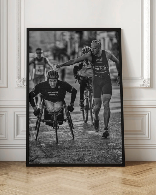 A dynamic black and white action photograph of a para-athlete in a racing wheelchair during a race. A running athlete beside him reaches out to hand him a sponge, showcasing a moment of camaraderie and support. Other competitors are blurred in the background on the wet asphalt. Print