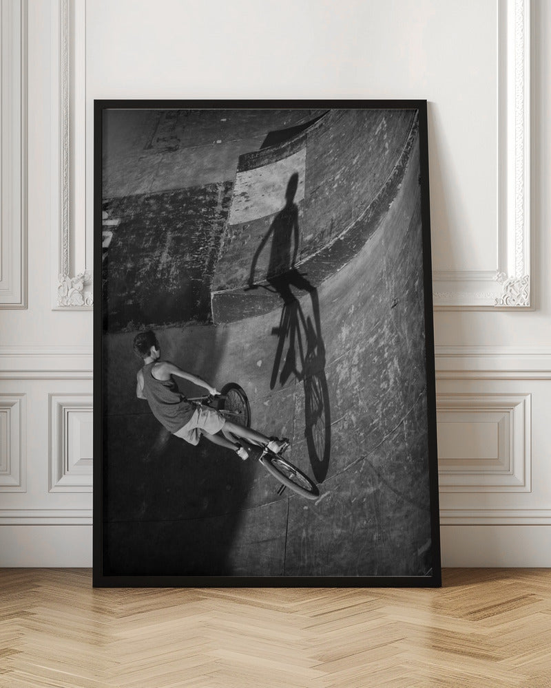 A dramatic black and white high-angle photograph of a BMX rider in a skate park. The rider is captured in motion on a curved ramp, while their long, stark shadow is cast on the worn, textured surface ahead, creating a powerful visual contrast. Poster