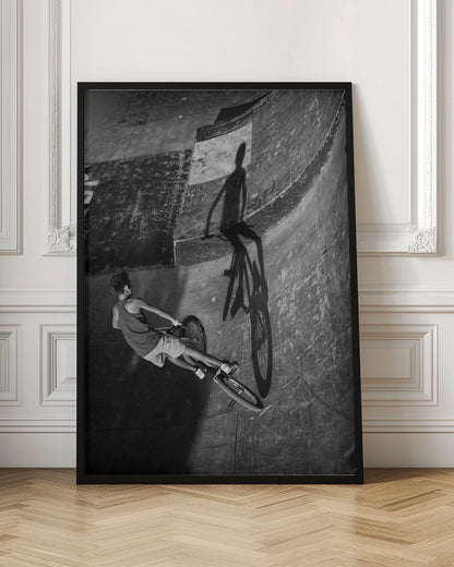 A dramatic black and white high-angle photograph of a BMX rider in a skate park. The rider is captured in motion on a curved ramp, while their long, stark shadow is cast on the worn, textured surface ahead, creating a powerful visual contrast. Poster