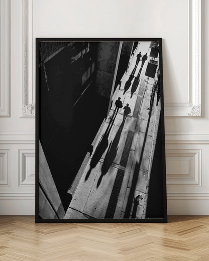 A dramatic high-angle, black and white photograph of several people walking on a sunlit sidewalk, casting long, stark shadows that stretch diagonally across the frame, contrasting with the deep black shadows of the adjacent building. Decor