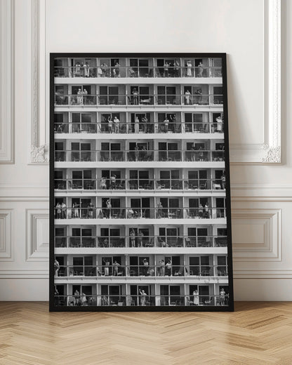 A black and white, high-contrast photograph of the side of a cruise ship, showing a grid of balconies across multiple decks. Various passengers are visible on their balconies, some waving, some relaxing, and some observing, creating a pattern of individual human stories within the repetitive architectural structure. Poster