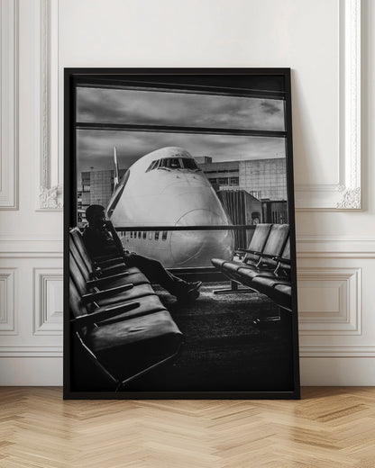 A dramatic black and white photograph of a silhouetted person sitting in an airport departure lounge, waiting for their flight. Through the large window, the enormous nose of a Boeing 747 jumbo jet is visible up close, dominating the view under a cloudy sky. Artwork