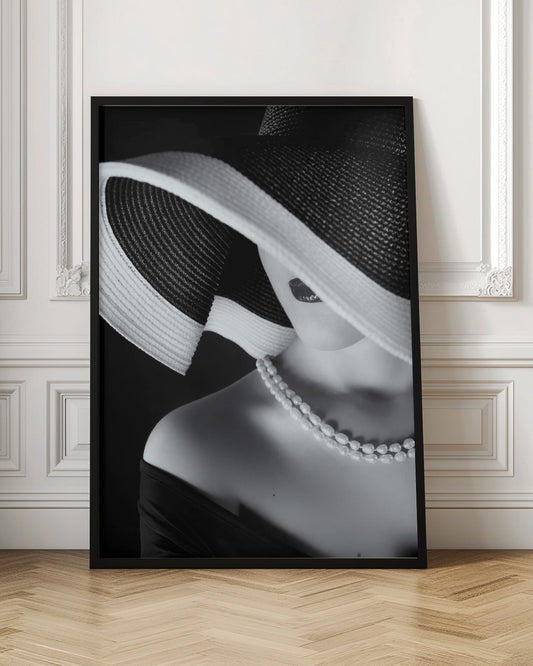 A glamorous black and white close-up photo of a woman wearing a large, striped sun hat that hides her eyes, revealing only her dark lips. She wears a pearl necklace and an off-the-shoulder top against a dark background, creating a mysterious and elegant mood. Wall Art