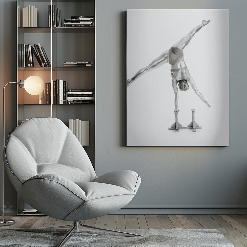 A black and white studio photograph of a muscular gymnast coated in silver paint, performing a dynamic one-armed handstand on a small block. Their legs are split wide apart in a display of strength and flexibility against a clean white background, all enclosed in a black frame. Decor