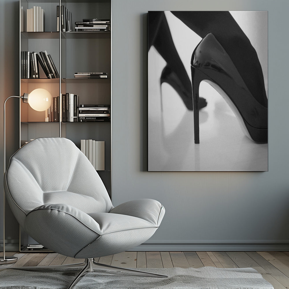 A close-up, black and white photograph of a person wearing shiny black patent leather stilettos and dark stockings. One shoe is in sharp focus in the foreground, highlighting its high arch and thin heel, while the other leg and shoe are softly blurred in the background on a reflective white surface. Decor