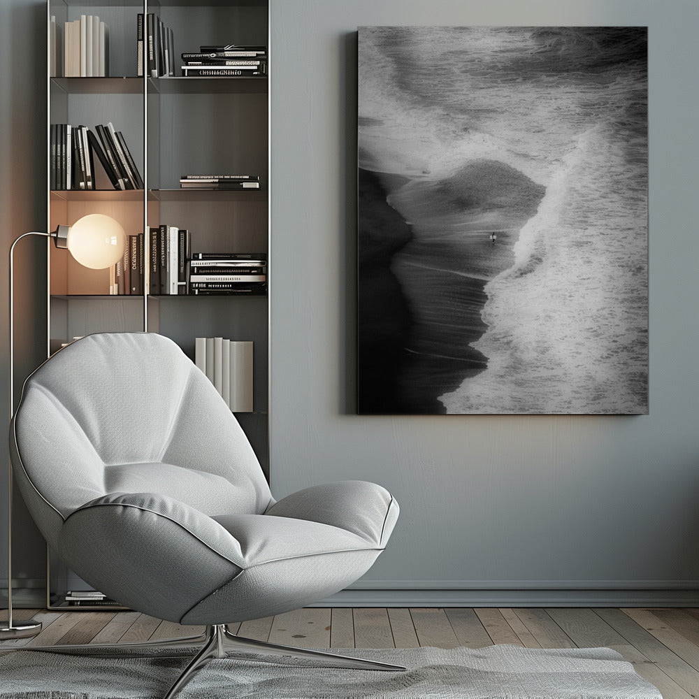 A high-angle, black and white photograph of a lone surfer carrying a surfboard at the water's edge on a dark sand beach. The vast, textured ocean with large, foamy waves crashing ashore dwarfs the solitary figure, creating a sense of scale and solitude. Decor