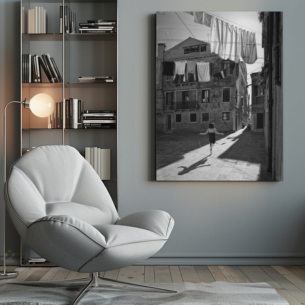 A black and white photograph capturing a girl from behind as she jump-ropes down a sunlit, narrow cobblestone street in Venice. Her long shadow is cast before her, and above, laundry hangs on clotheslines strung between old, weathered European buildings. Wall Art