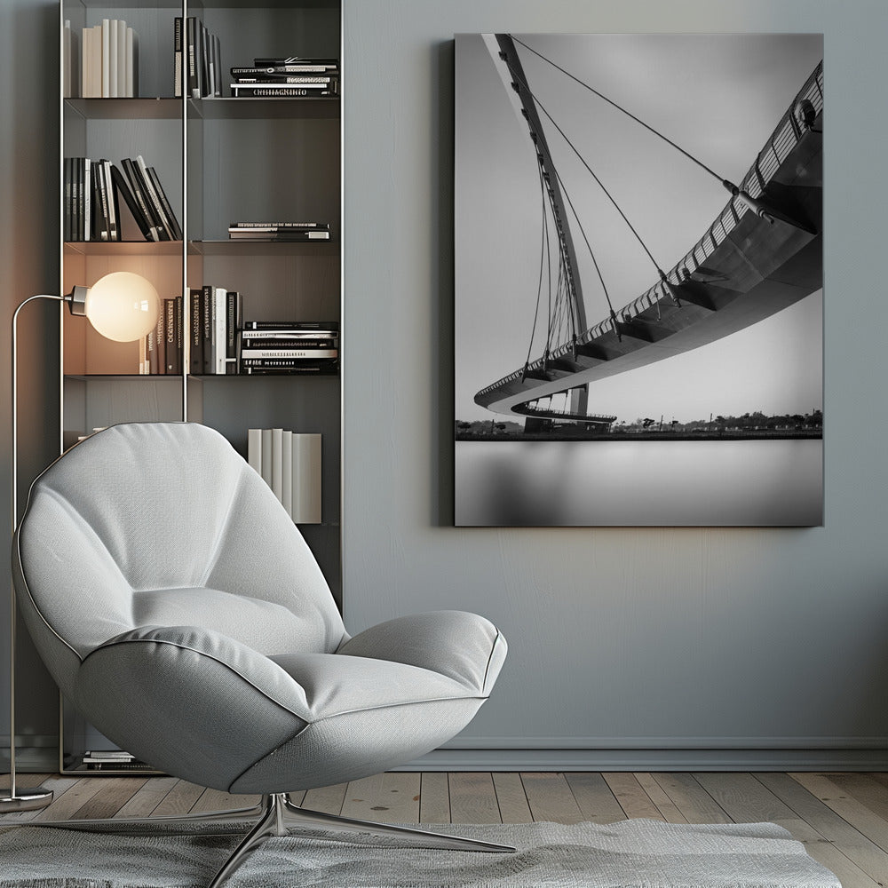 A dynamic low-angle, black and white photograph of a modern cable-stayed pedestrian bridge. The bridge curves elegantly over calm, reflective water, with its deck suspended by numerous cables attached to a tall, angled pylon, creating a striking graphic composition against a bright, clear sky. Decor