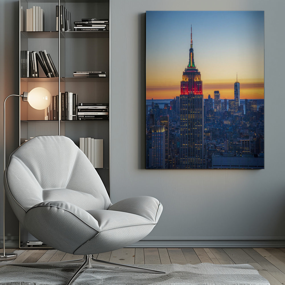 A vertical photograph of the New York City skyline at sunset, with the Empire State Building in the foreground. The building is illuminated with festive red and green lights on its upper floors, set against a sky that transitions from deep blue to a warm orange glow at the horizon. The surrounding cityscape, including the distant One World Trade Center, is bathed in the cool light of dusk, with scattered warm lights from building windows. Decor