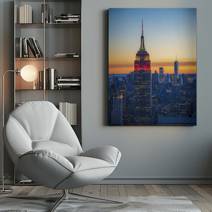A vertical photograph of the New York City skyline at sunset, with the Empire State Building in the foreground. The building is illuminated with festive red and green lights on its upper floors, set against a sky that transitions from deep blue to a warm orange glow at the horizon. The surrounding cityscape, including the distant One World Trade Center, is bathed in the cool light of dusk, with scattered warm lights from building windows. Decor