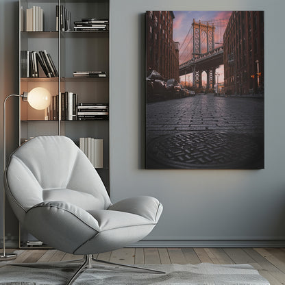 A low-angle photograph taken from a cobblestone street in Dumbo, Brooklyn, looking up towards the Manhattan Bridge at sunset. Red brick buildings frame the view on both sides of the street, with parked cars lining the left. A manhole cover is prominent in the foreground, and the sky is filled with warm pink and orange tones. Print