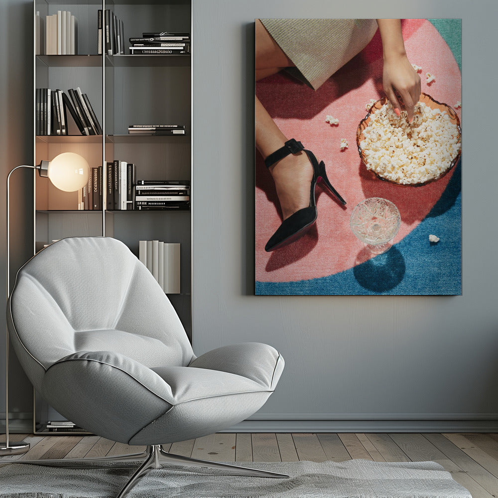 A high-angle photograph of a woman sitting on a vibrant pink and blue rug. She is wearing a chic black stiletto high heel and reaching into a glass bowl filled with popcorn. A crystal glass and scattered popcorn kernels are also on the rug. Artwork
