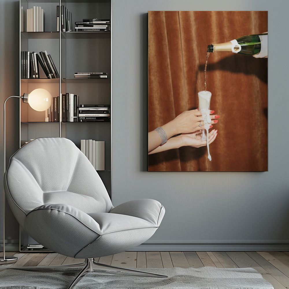 Champagne is poured from a bottle into a glass, overflowing with white foam that drips down onto two manicured hands with red nail polish holding it against a brown velvet curtain background. Poster