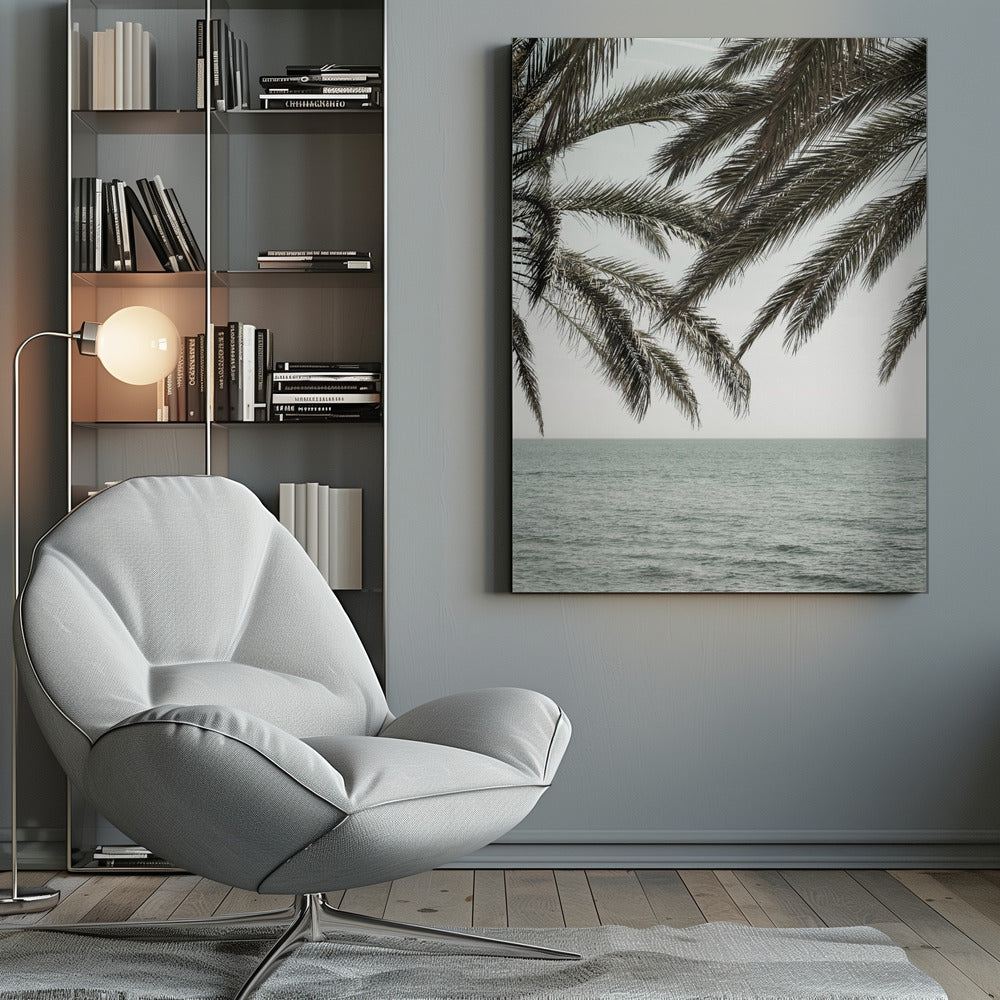 A close-up view of dark green palm tree fronds hanging over a calm, muted sage green ocean under a pale, overcast sky. The photograph has a serene and tranquil mood and is displayed in a black frame. Wall Art