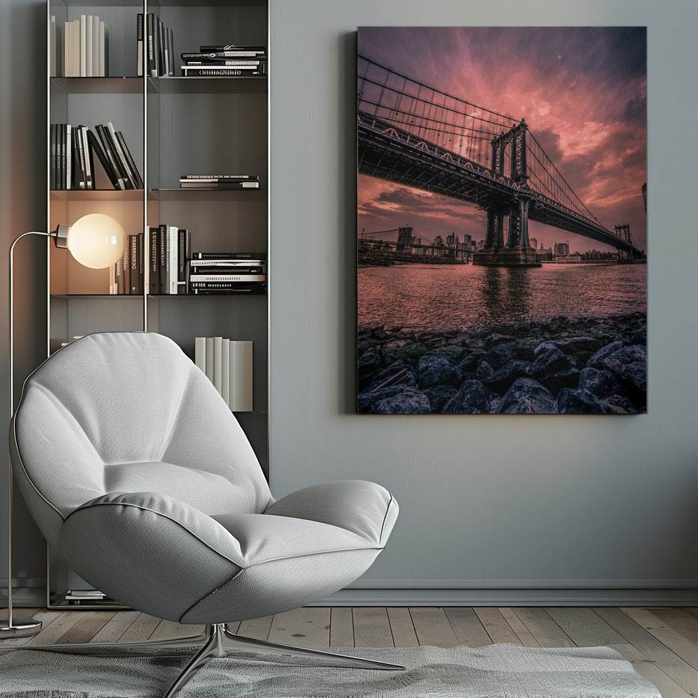 A dramatic low-angle view of the Manhattan Bridge over the East River at sunset. The sky is filled with pink and red clouds, which are reflected in the water below. The dark silhouette of the bridge contrasts with the vibrant sky, and the New York City skyline is visible in the background. In the foreground, dark, wet rocks line the riverbank. Decor