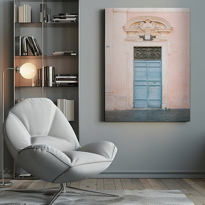 A light blue, studded double door set into a pale pink wall. The doorway is framed in white stone with an ornate pediment and a decorative wrought-iron transom window above the door. A single pigeon stands on the cobblestone pavement in front. Print